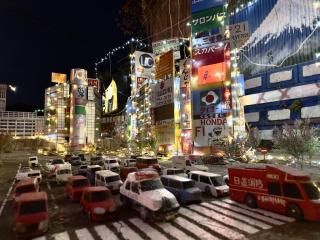 Outdoors at night, a model version of shibuya with wooden carved cars in front and buildings lit up with strings of LEDs in back, with Shibuya 109 at the far rear. The signs are all hand-drawn.