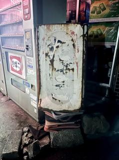 A sun-faded old sign where you can barely make out たばこ which is posted ontop of old cracked traffic cones with a running cigarette vending machine in the background 