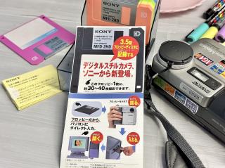 A pack of various colored floppies, a Mavica digital camera and a flyer that says3.5型フロッピーディスクに(DOS/Vフォーマット）記録するデジタルスチルカメラ、ソニーから新登場。このフロッピー1枚に、約30〜40枚撮影できます。フロッピーだから、パソコンにダイレクト入力。