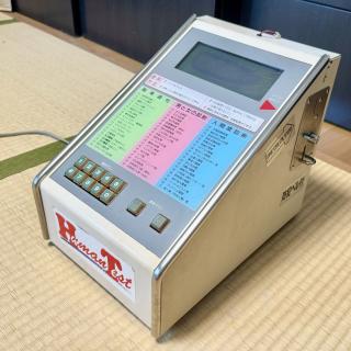 A device that says “Human Test” on the front, has 3 columns of Japanese text with numbers, and a 4-line black and white LCD display. On the side is a coin slot.