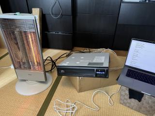 A 2U rackmount APC UPS lying on top of (and crushing) the shipping box it came in, while hooked up to a glowing IR radiant space heater, while a laptop shows the state of charge