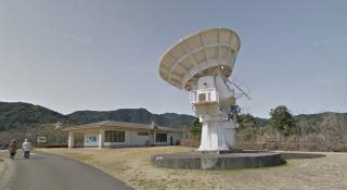 A decent sized white satellite dish on a concrete pedestal in a grass field. You can make out the blue information panels from the first post on a building in the background