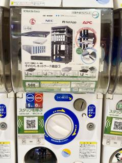 A gacha machine showing the contents being a network rack with various known brand names like netapp and NEC