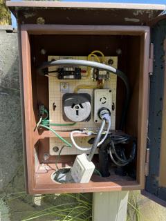 An outdoors power box, with visible wiring and a breaker and two different kinds of outlets, one large one that looks like an Australian plug but hecking chonker and one smaller one that has three circular pins. The ground block is quite corroded.Into the skinny outlet is connected a clearly hand-made cheater plug which has a standard wall outlet stuck on the end