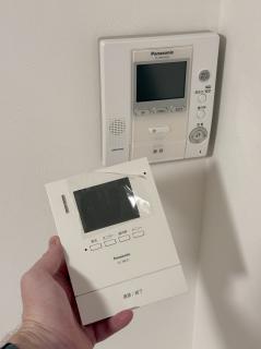 A Panasonic VL-ME31 door intercom with the plastic film still on the display, held in front of a VL-MW102K that is only very slightly yellowed on the middle plastic piece.