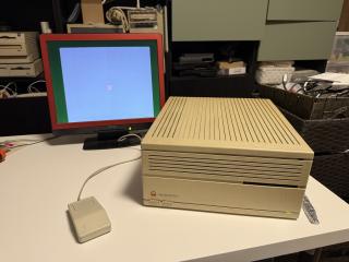 An old beige Macintosh IIcx with a rainbow Apple logo on a desk with only a mouse plugged in and an LCD monitor with a floppy disk icon on it