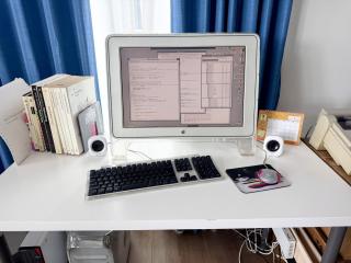 A desk with an Apple Cinema Display, Apple Pro Keyboard, pink iMac puck mouse and a bookstand with a series of Inside Macintosh books. Beneath the desk a PowerMac G4 can be glimpsed