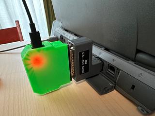 The rear of a PowerBook 540c showing the ports, with a SCSI HDI adapter in the back with a green 3D-printed device plugged in, and a USB-C cable coming in the top. Inside the green case a red LED is shining