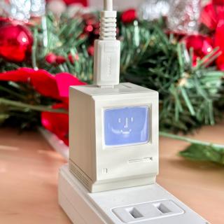 What looks like a Classic Mac with a pixelated Happy Mac face that is lit up in the kind of bluish-white of a Classic Mac CRT. But at a second glance it's plugged into a power strip and you realize it's tiny. There's a beige USB-C cable with an old-fashioned looking strain release plugged into the top, since this is a USB-C charger.
