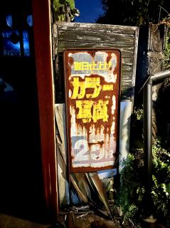 A rusty old sign with rotted wood behind it and plants growing around that says 翌日仕上げカラー写真25円