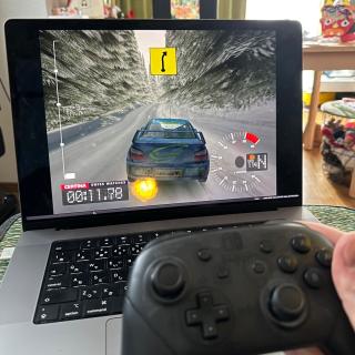A MacBook Pro playing Colin McRae Rally 3 for the PS2 under emulation, with a hand holding a Nintendo Switch Pro controller in the foreground.
