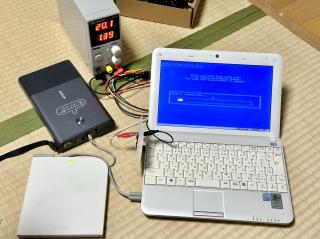A small white laptop running the Windows XP installer hooked up to a bench power supply with a USB optical drive attached with is also attached to a massive Anker battery