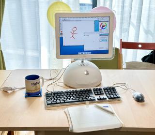 An iMac G4 with a Wacom tablet and pen in front of it. On the screen is Corel Painter 8 and MacOS X Ink.SPOLIER:(For the eagle-eyed viewer, off to the corner there is a grounded cable plugged into an extension cord in a way that the grounded pin sticks out)