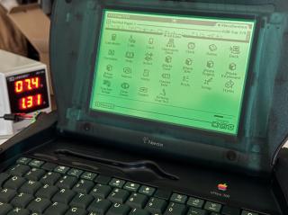 An Apple eMate 300 indicating that it is charging the internal battery. In the rear a bench power supply is seen indicating 1.3A of draw