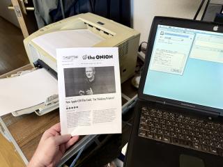 A Laser Printer and a PowerBook G3. A hand is holding a printout of an article from The Onion with a photo of Tim Cook and the headline “New Apple CEO Tim Cook: I'm Thinking Printers”
