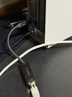 A small metal box with PLANEXCOMM, USB-C and 2.5Gbps written on it with an Ethernet cable going out one end and a USB-C going out the other. It is plugged into a small black USB-C to A adapter going into a PC next to the PS/2 mouse and keyboard ports.