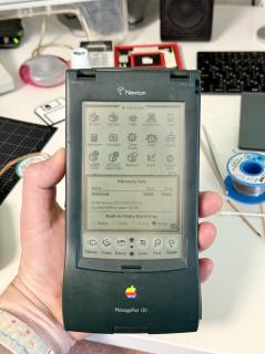 A Newton MessagePad that is on and showing a memory info screen with 1024K of memory and Newton OS 2. In the background is a messy desk full of soldering equipment