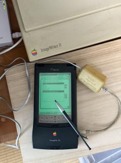 A Newton MessagePad 130 showing a long AppleTalk Zones list, connected to a PhoneNET PLUS adapter. An ImageWriter II and AsanteTalk are also visible in frame