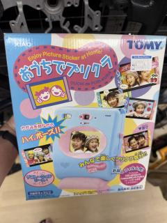 A box with a photo of something that resembles an instax camera but has what appears to be a screen in the middle. The box is clearly from the early 2000's though. On closer inspection, the lens and flash are on the front??TOMY.Enjoy Picture Sticker at Home!もうちでブリターOUCHI de PURI-KURAかがみを見ながらハイボーズ！！