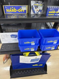 A shelf of merch with the HARD OFF blue color and logoStickersRubber keychainAlkaline batteriesMini “Junk” traysCarry bag