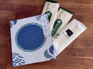 A box with a blue Japanese style pattern on it with what appears to be a seeing eye image, and the text 恩返し enひかり. Under it are three packs of soumen noodles