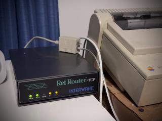 The router box on a desk with its activity lights lit next to an ImageWriter II.